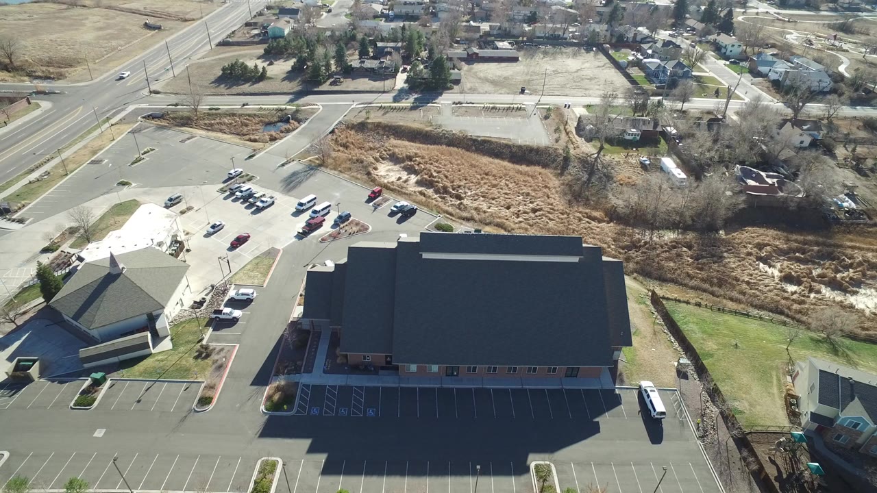 High aerial view in Hidden Lake, Colorado showing two large dark-roofed buildings, paved parking lots, nearby roads, dry grass, and no visible people.