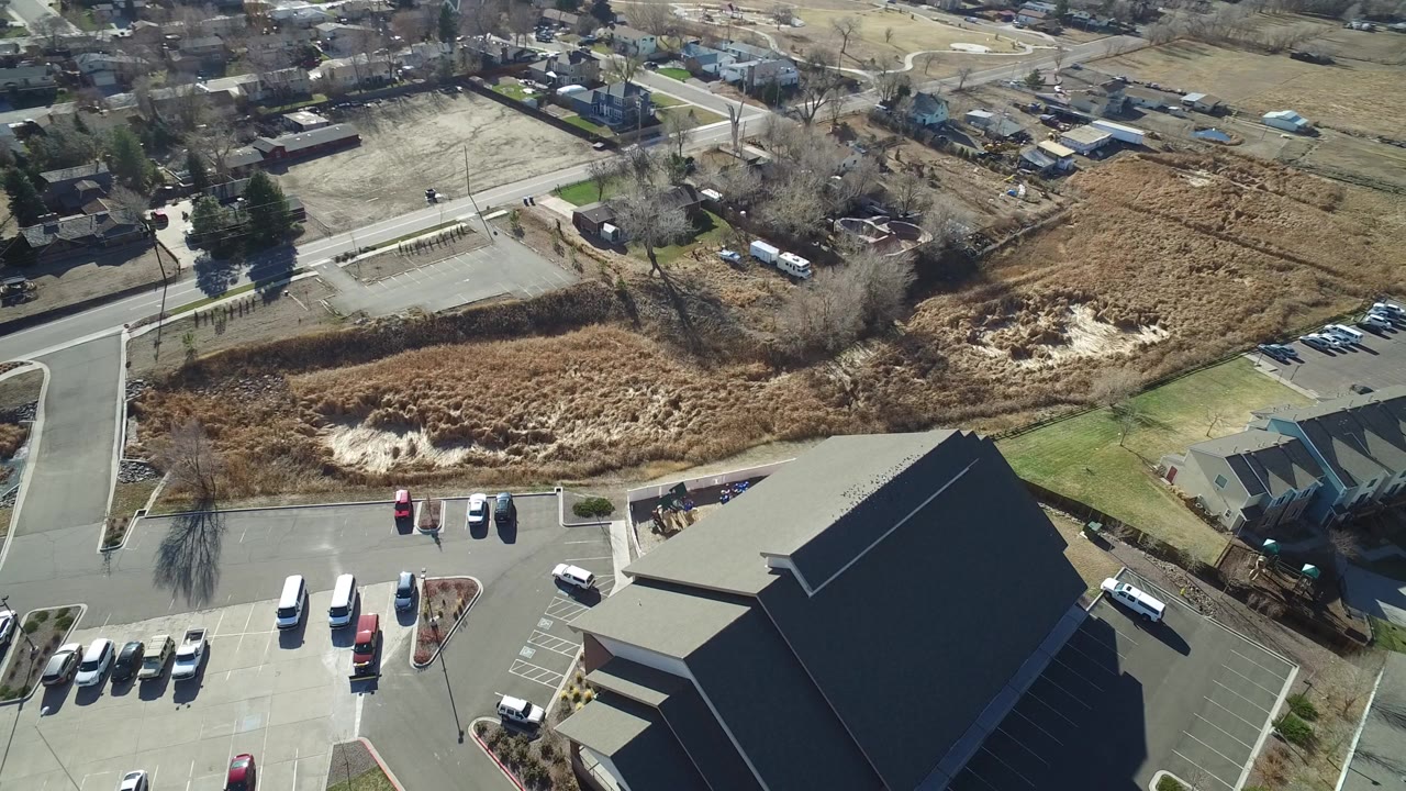 Aerial view of parking lots, a dark-roofed building, roads, homes, and dry open land.