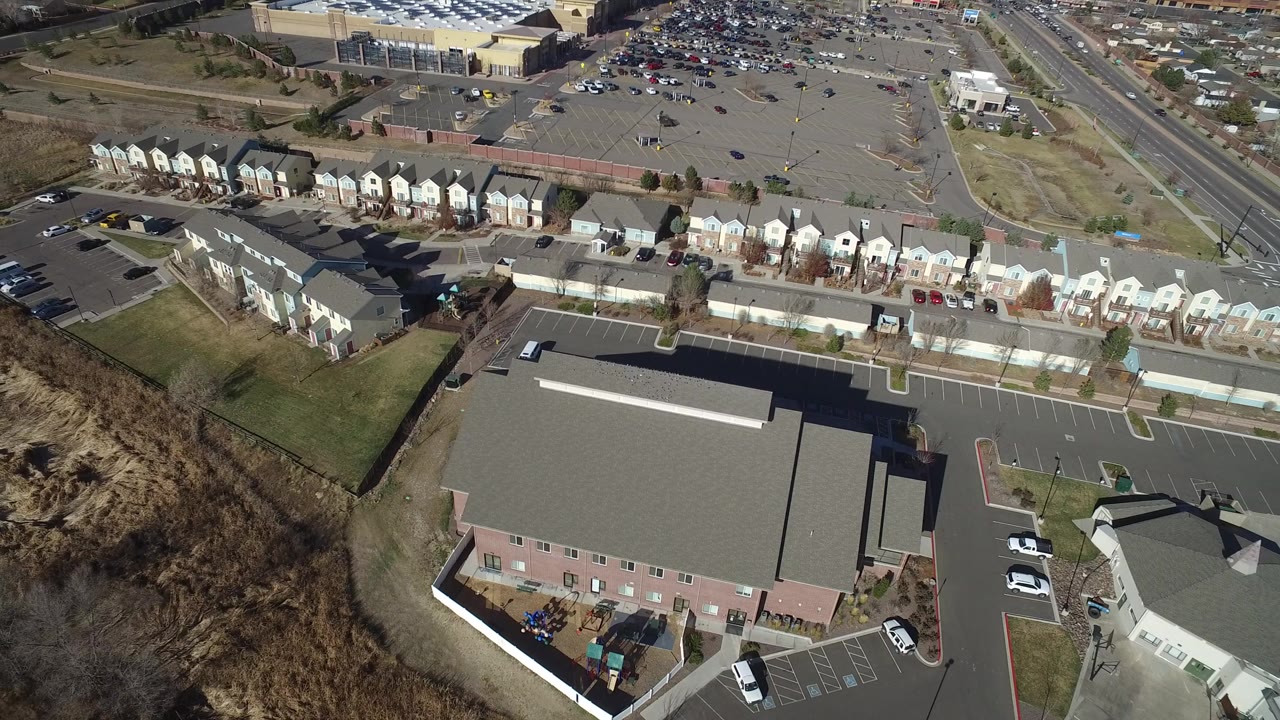 Overhead view of a large building, parking lots, townhomes, roads, and parked cars.
