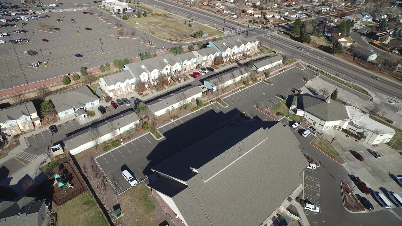 Top-down aerial frame over Hidden Lake, Colorado showing a large roofed building in the lower foreground, rows of homes, roads, parking lots, cars, dry grass, and no visible people.