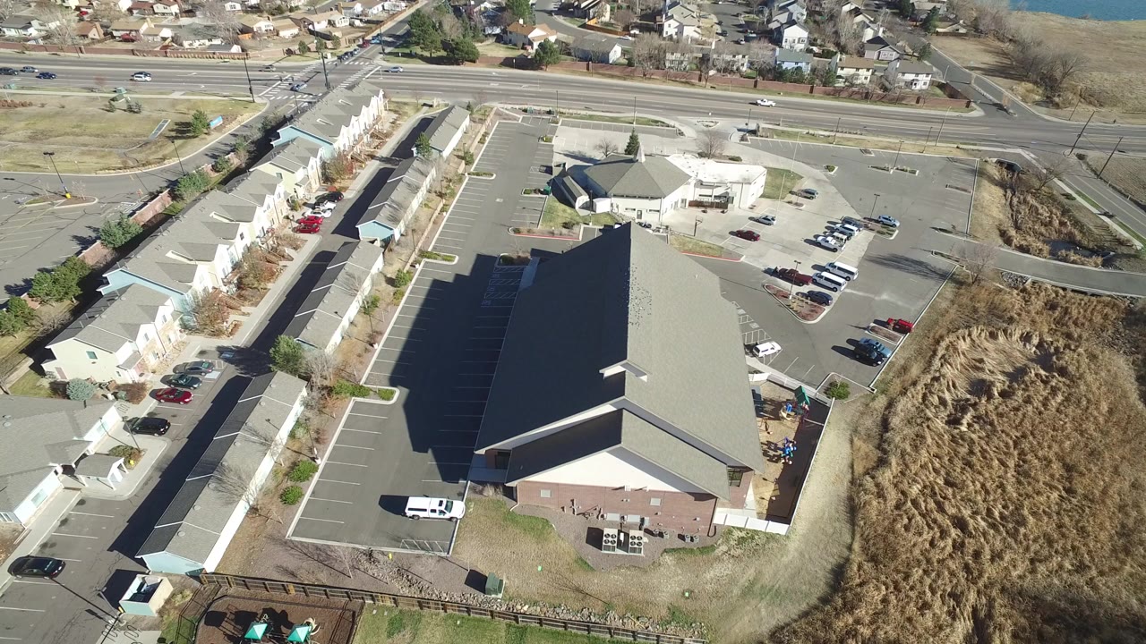 Aerial view of a large gray-roofed building, parking lots, roads, and nearby homes.