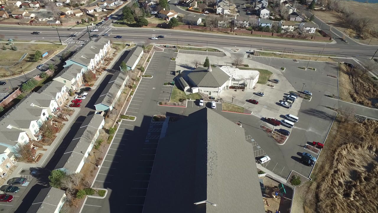 High aerial view in Hidden Lake, Colorado showing a large parking lot, a hexagonal-roofed building, attached residential buildings, a road intersection, and no visible people.