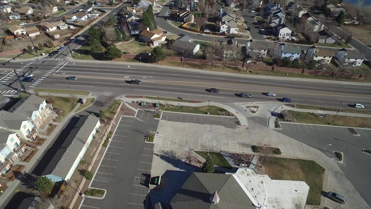 Aerial view in Hidden Lake, Colorado with cars spaced along a multilane road, a large mostly empty parking lot below, and residential roofs beyond, no visible people.
