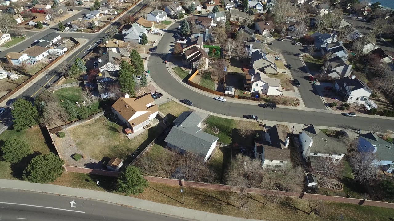 High aerial view in Hidden Lake, Colorado with detached homes around a curved street, a larger road in the foreground, and no visible people.