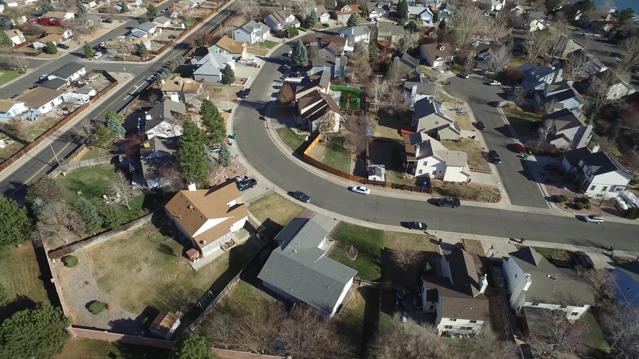 Aerial neighborhood block with rooftops, yards, and several cars.