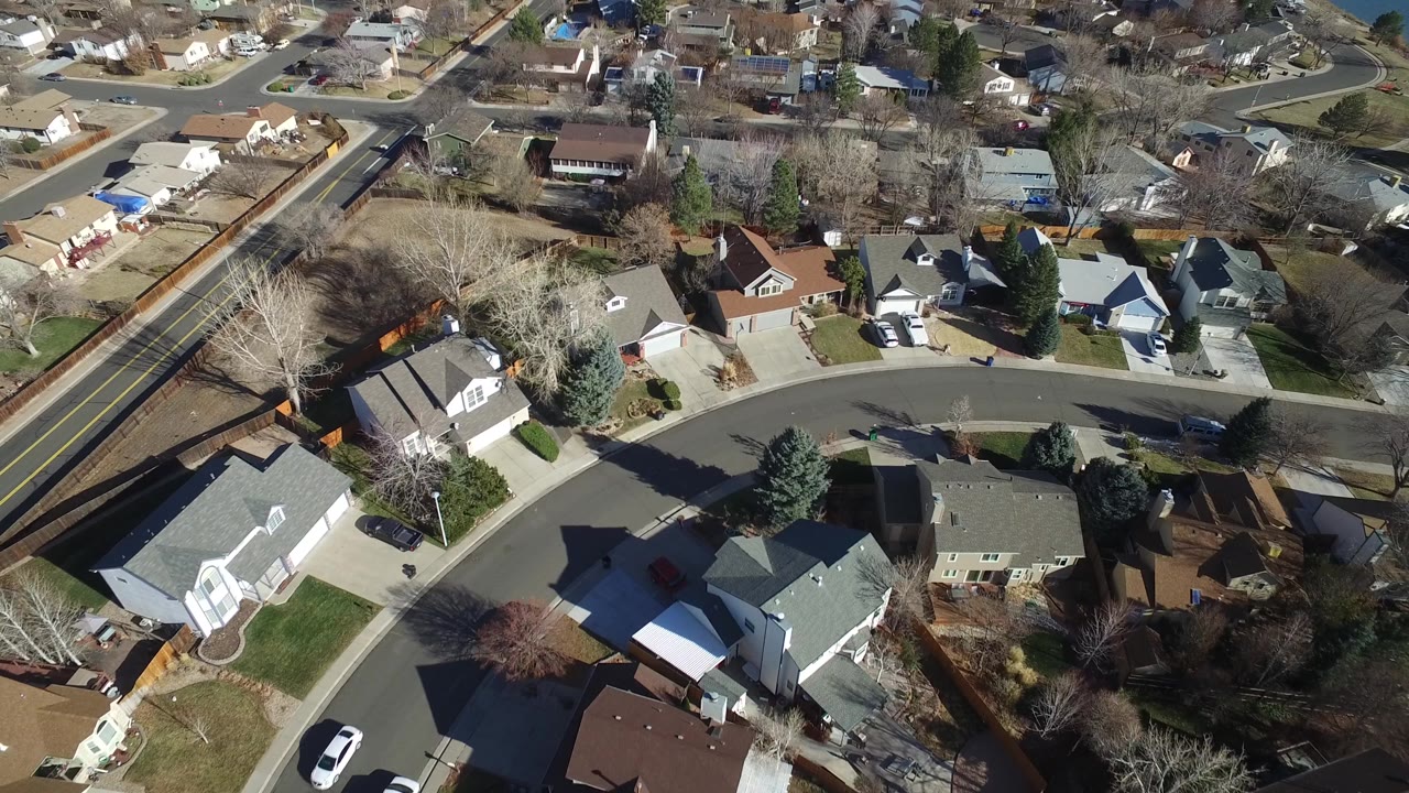 The second frame looks down across Hidden Lake, Colorado homes, backyard fences, a two-lane road near the top, parked vehicles, and winter-bare trees, with no visible people.