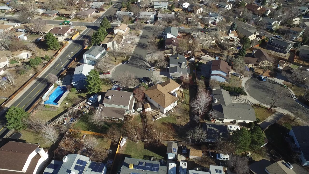 Opening aerial frame over Hidden Lake, Colorado homes with a two-lane road at left, fenced backyards, parked vehicles, leafless trees, and no visible people.
