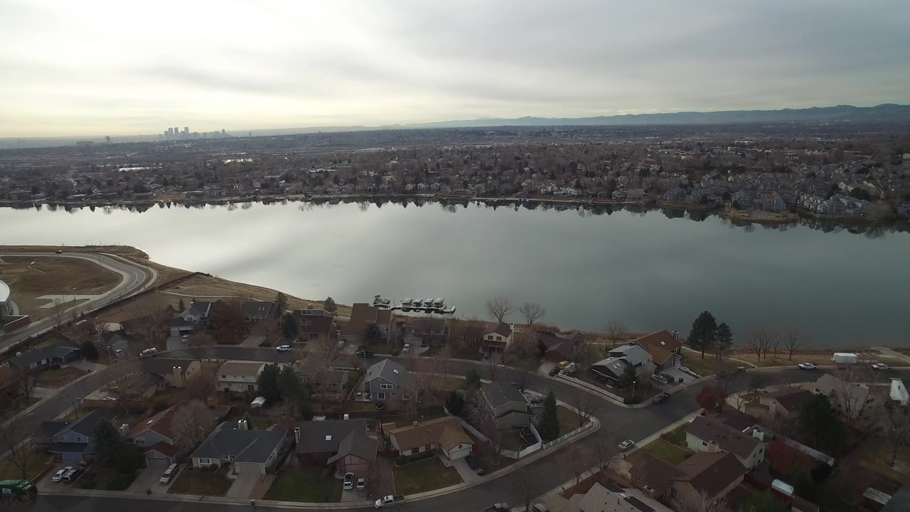 Aerial frame in Westminster, Colorado looking across calm water toward dense suburbs, distant city buildings, low hills, and no visible people.