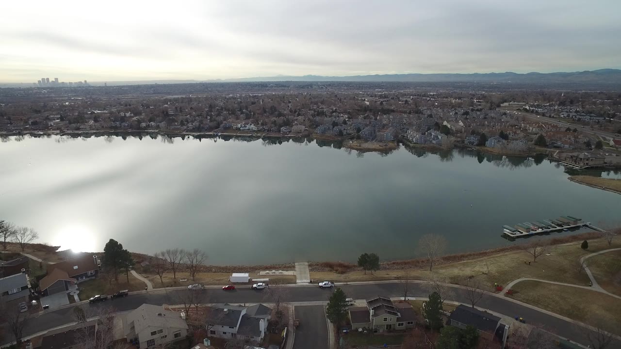 Aerial view over a Westminster, Colorado lake with a shoreline road, nearby houses, docks to the right, distant buildings, mountains, gray clouds, and no visible people.