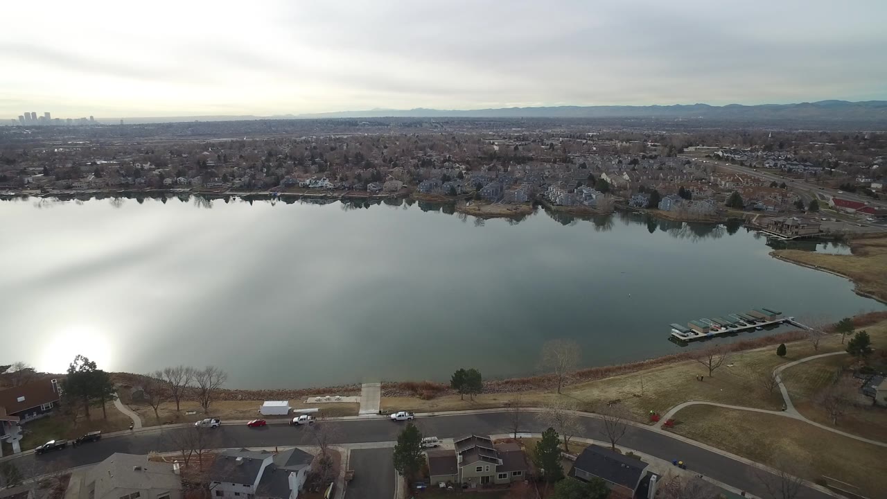 Drone view across a lake toward suburban homes, docks, roads, and mountains in the distance.