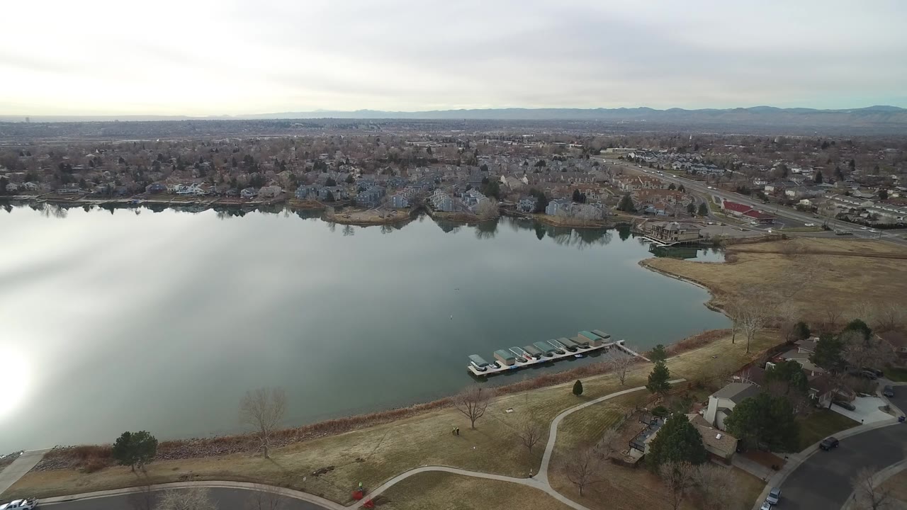 Aerial view across the Westminster, Colorado shoreline, with covered boats on a dock in the foreground, houses near the water, roads beyond, muted grass, and no visible people.
