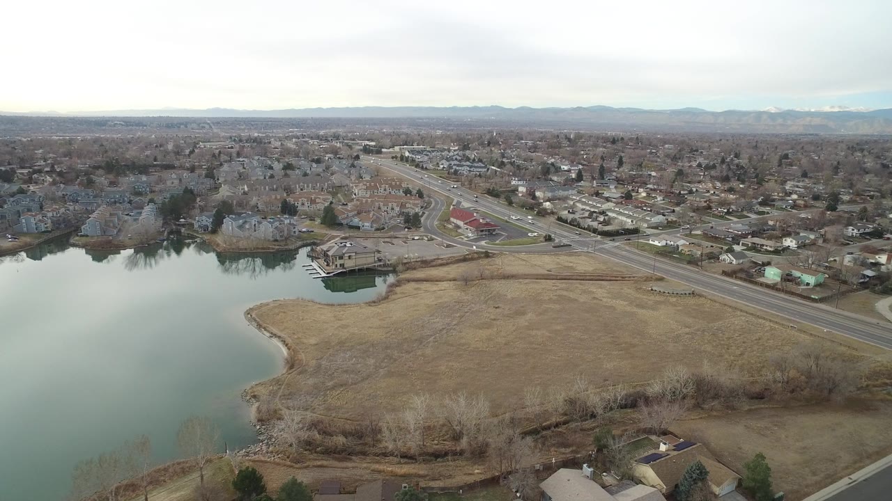 High aerial frame over Westminster, Colorado showing a lake at left, dry grassland in front, a road crossing the scene, residential blocks beyond, distant mountains, and no visible people.