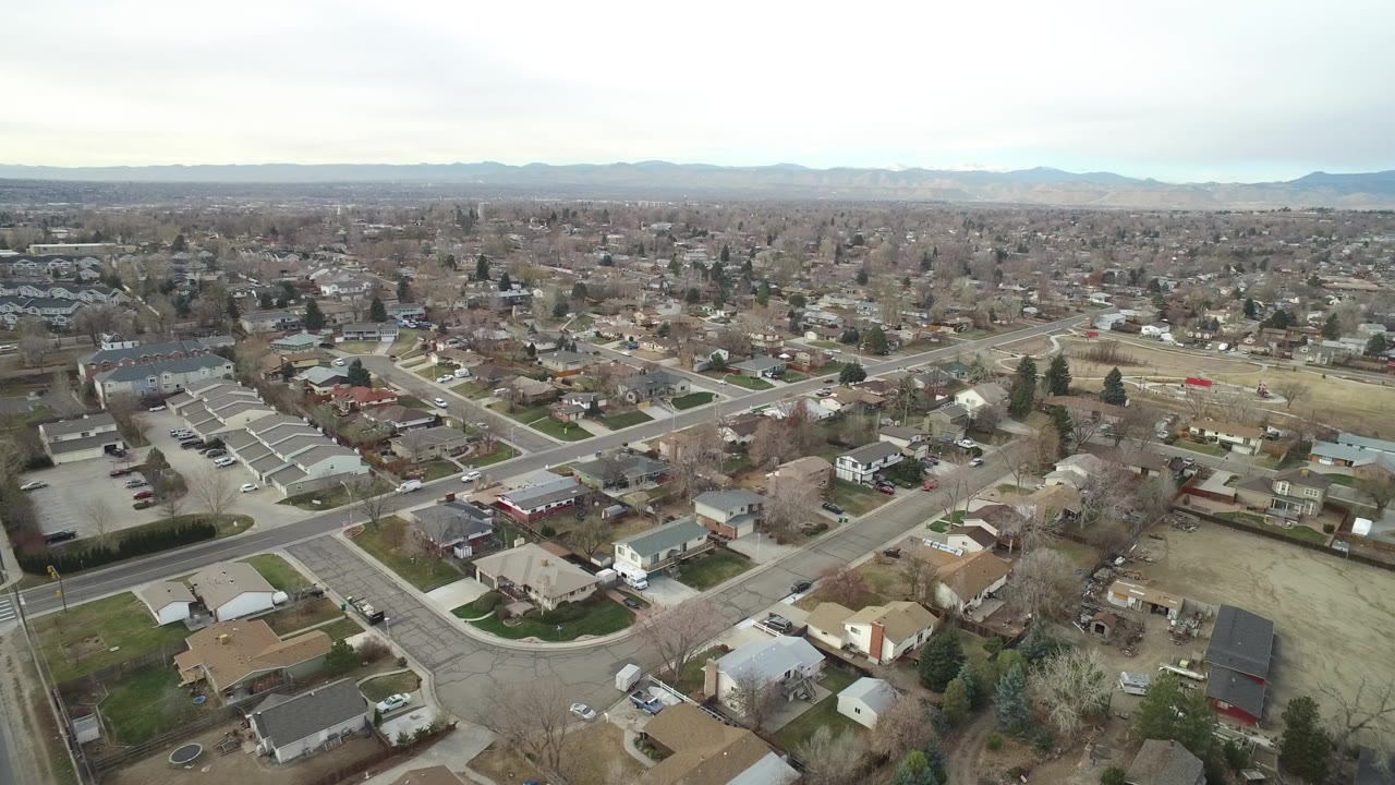 A high aerial frame over Westminster, Colorado shows residential streets, single-family houses, townhomes, bare trees, parked cars, pale lawns, and distant mountains, with no visible people.