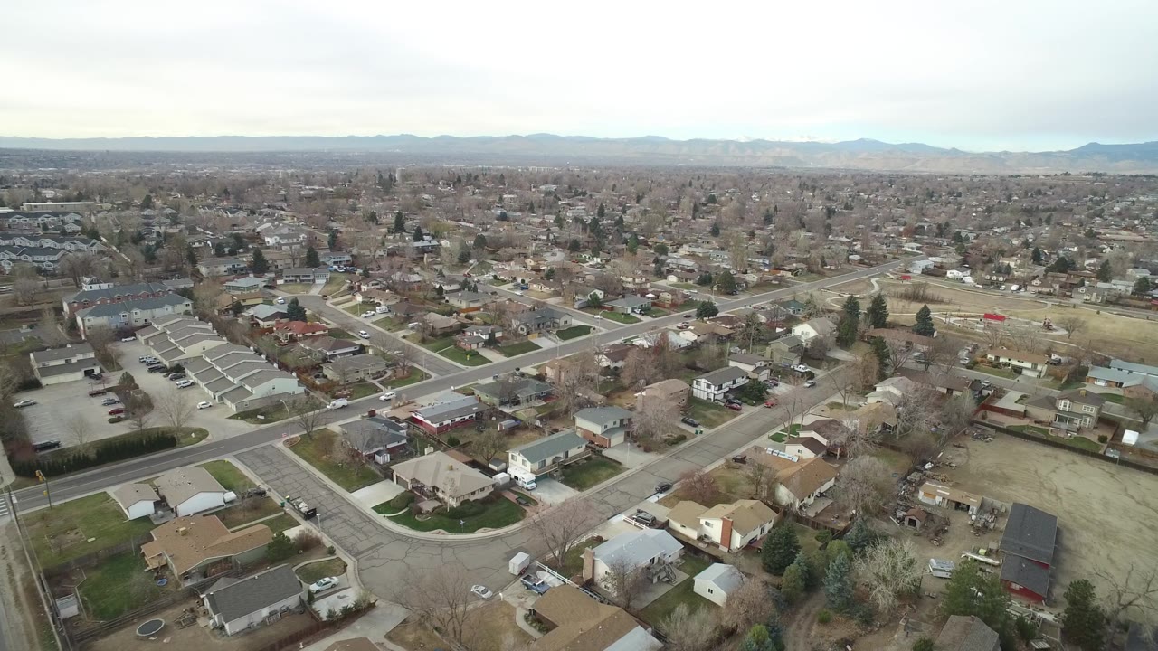 A wide aerial view of Westminster, Colorado shows an intersection and surrounding homes in the foreground, rows of residential blocks beyond, and a low mountain range under gray cloud cover, with no visible people.
