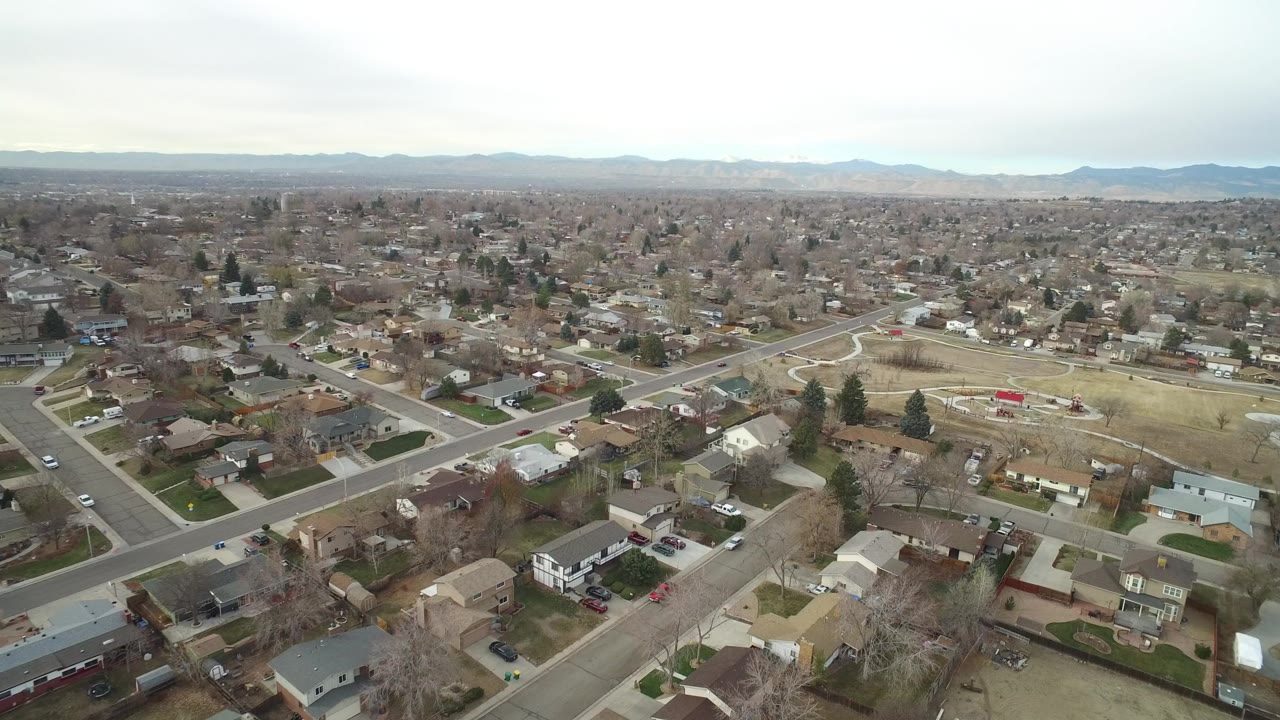 Opening aerial frame over Westminster, Colorado homes and neighborhood streets, with a small park and distant mountains visible under cloud cover, no visible people.