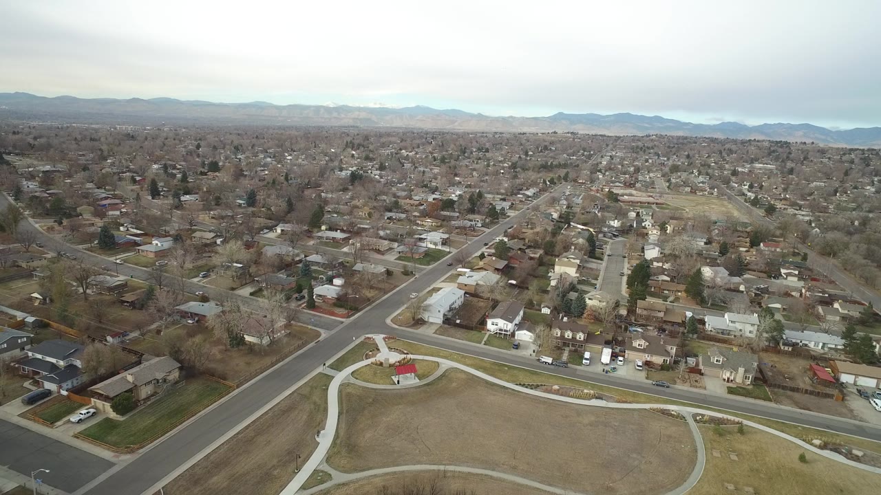 Elevated Westminster, Colorado view with a wide street crossing the foreground, rows of low houses, leafless trees, scattered vehicles, distant mountains, gray clouds, and no visible people.