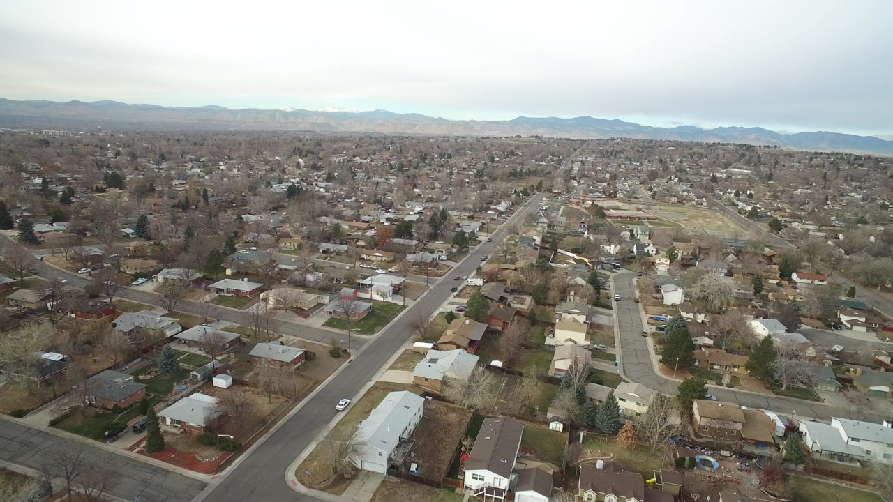 Aerial view of suburban homes, streets, trees, and distant mountains under clouds.