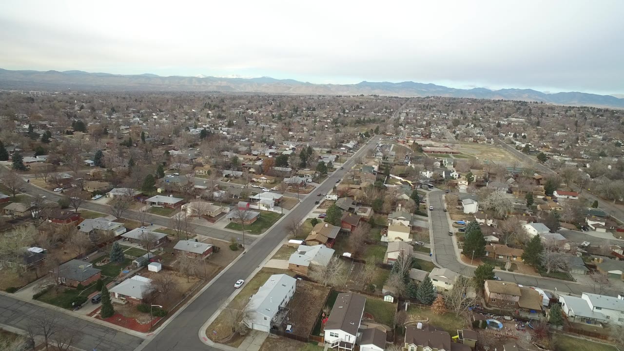 Aerial view in Westminster, Colorado, centered on a straight road running through houses, bare trees, muted lawns, and distant mountains, with no visible people.
