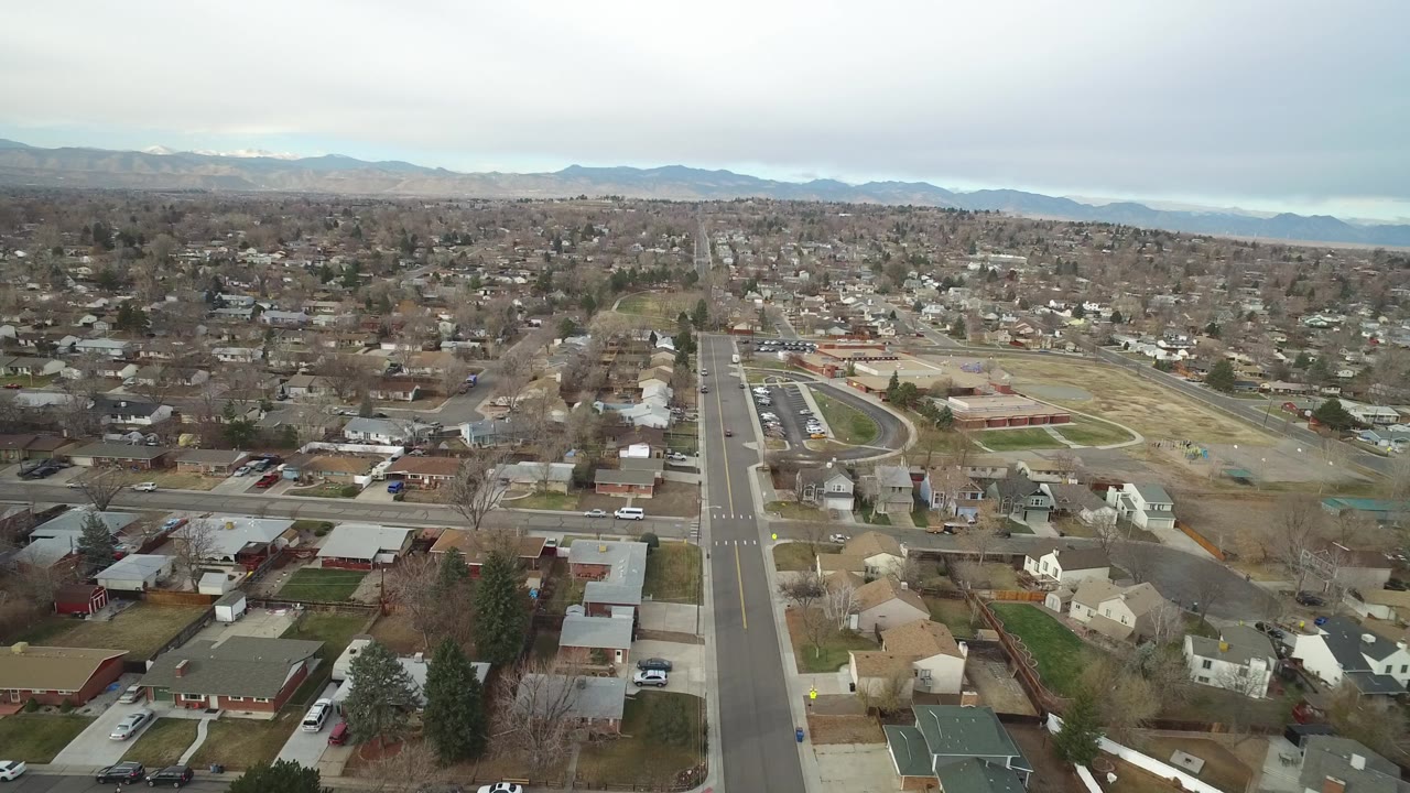 Aerial frame over Westminster, Colorado showing a central roadway, clusters of single-family homes, a field complex to the right, distant mountains, overcast light, and no visible people.