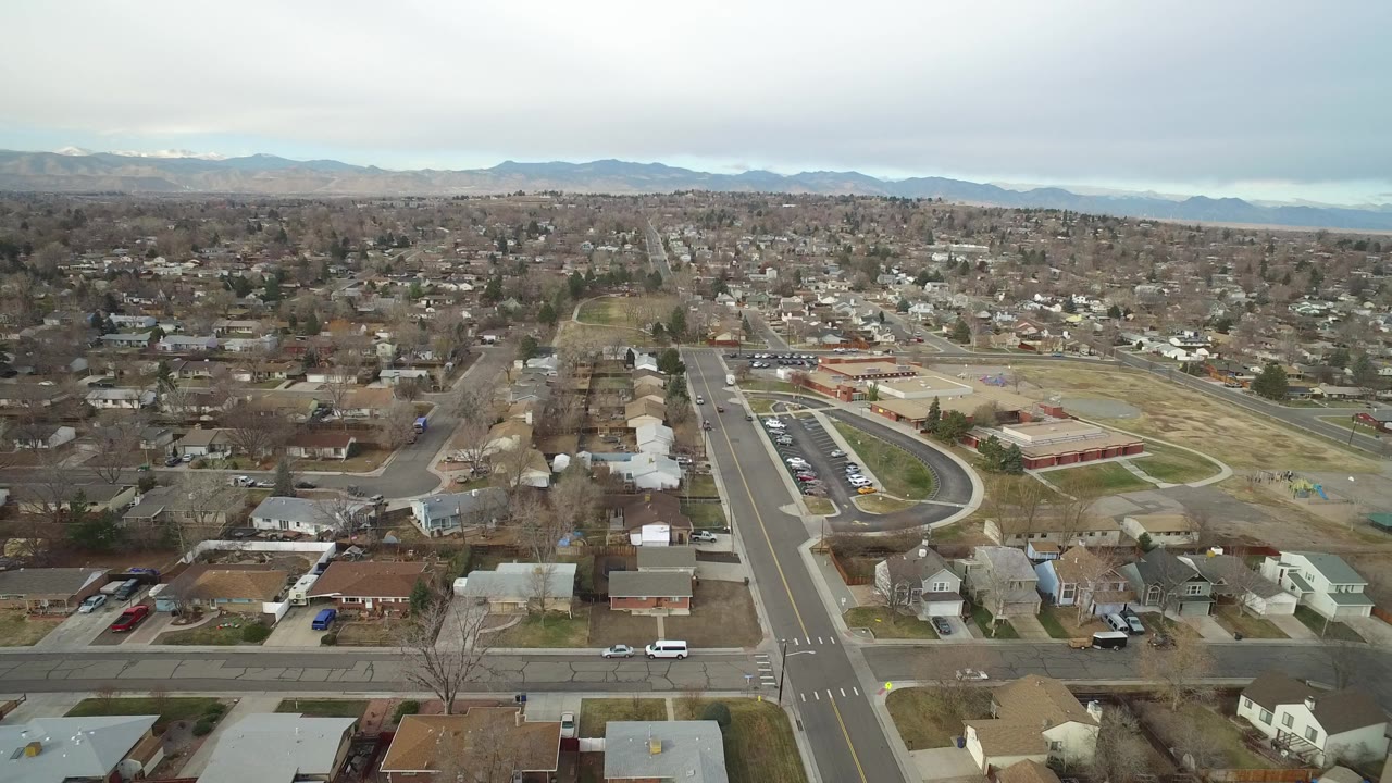 Opening aerial frame over Westminster, Colorado showing a broad intersection, residential blocks, a low public-looking building, muted winter trees, distant mountains, and no visible people.
