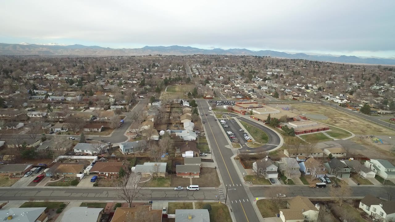 Bird's-eye suburban view with an intersection, houses, parking lots, open grounds, and mountains on the horizon.