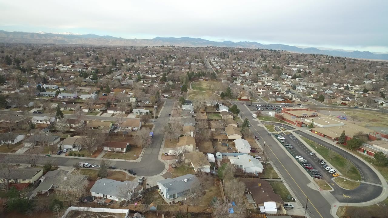 Aerial Westminster, Colorado frame with rooftops in the foreground, a road and parking area near a low building, mountains on the horizon, and no visible people.