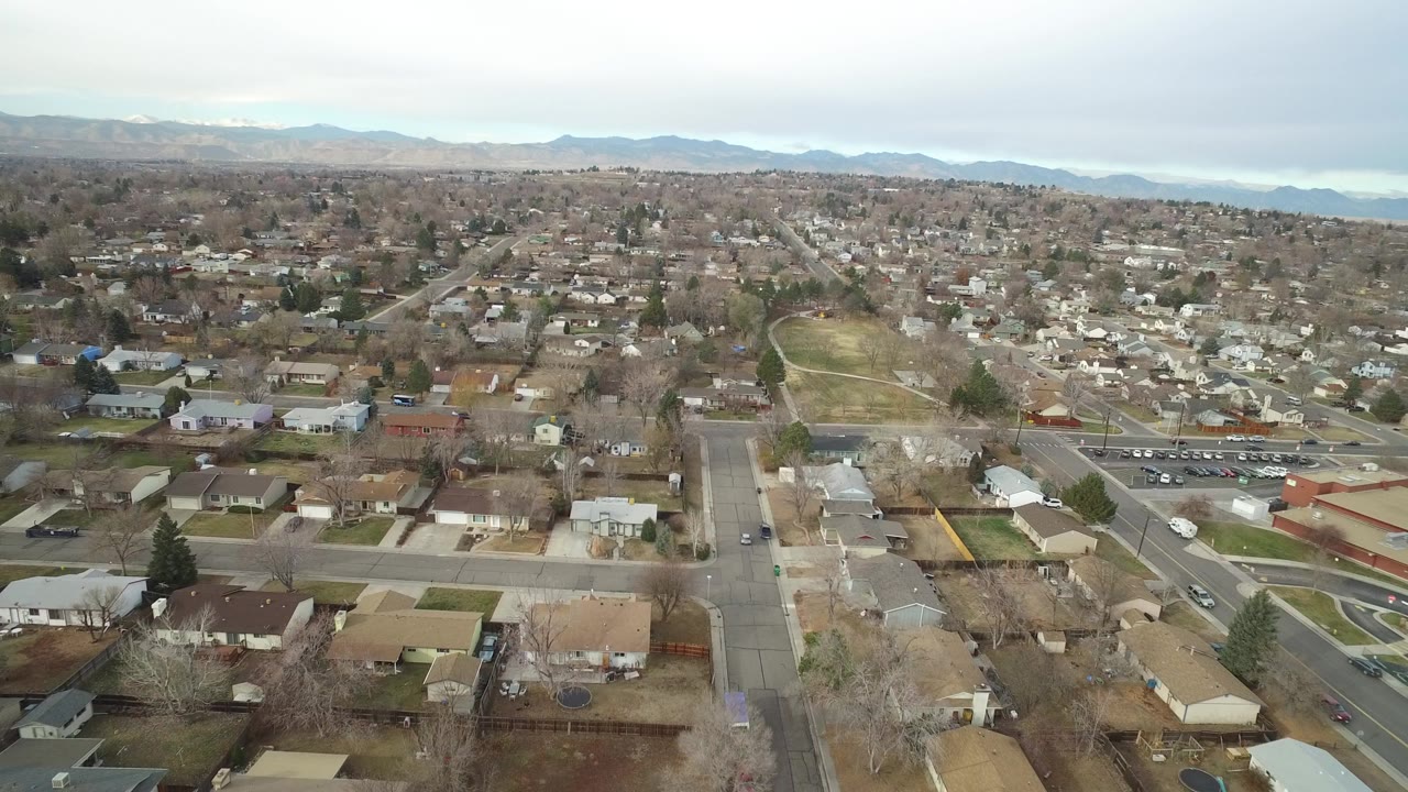 High aerial view over Westminster, Colorado homes and neighborhood streets with parked cars, bare trees, distant low mountains, overcast sky, and no visible people.