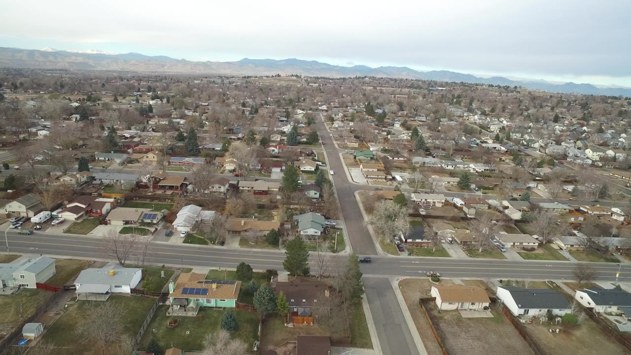 Wide drone view over Westminster, Colorado residential blocks, a main road crossing the foreground, distant mountains, overcast sky, and no visible people.