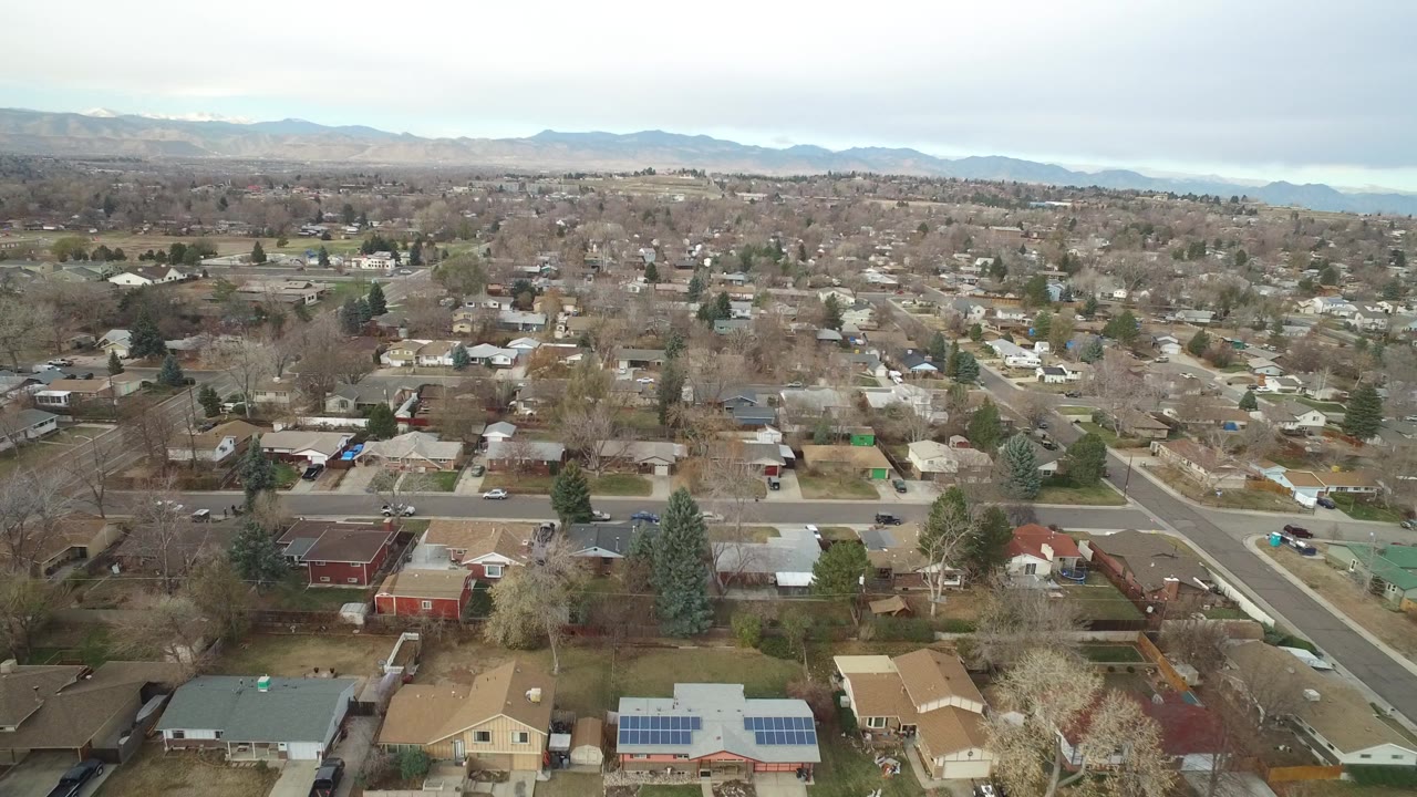 Opening aerial frame over Westminster, Colorado homes, tree-lined streets, parked cars, distant mountains, pale sky, and no visible people.