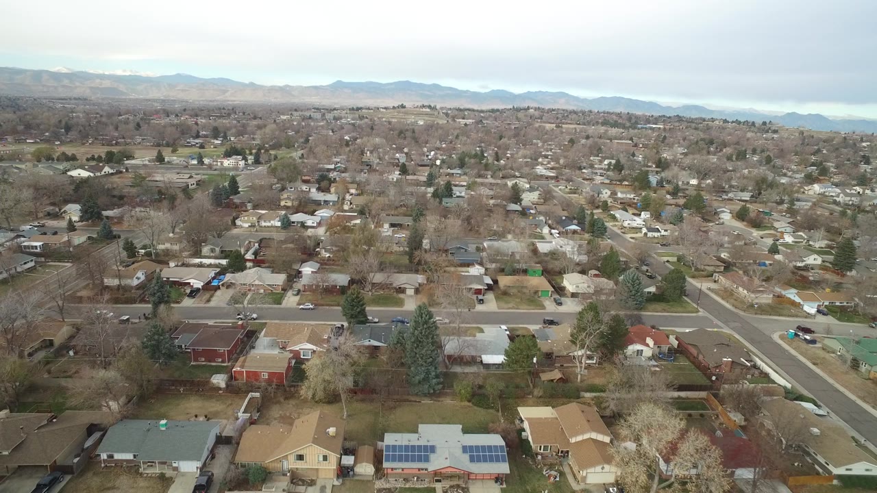 Aerial frame over Westminster, Colorado residential blocks with a central road, leafless trees, rooftops, distant foothills, and no visible people.
