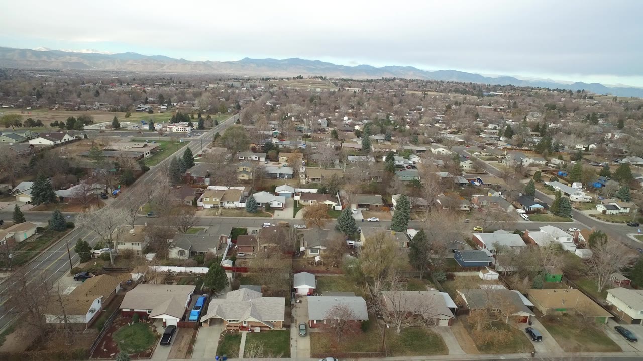 Aerial frame over Westminster, Colorado rooftops and bare trees, with a main road at left and a hazy mountain horizon, no visible people.