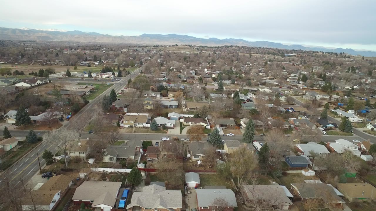 Wide Westminster, Colorado aerial with foreground houses, a diagonal arterial road, pale winter trees, and distant foothills beneath gray cloud, no visible people.