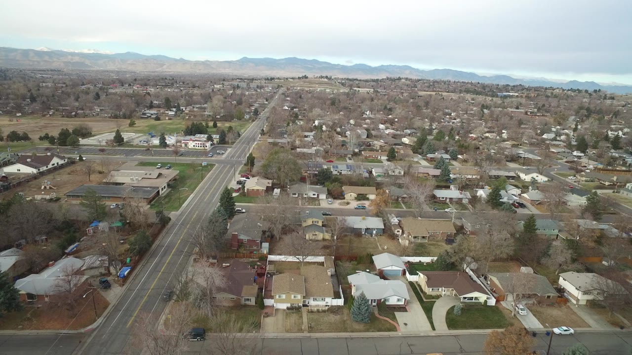 Early aerial frame over Westminster, Colorado showing residential roofs, a long diagonal roadway, bare trees, distant mountains, and no visible people.