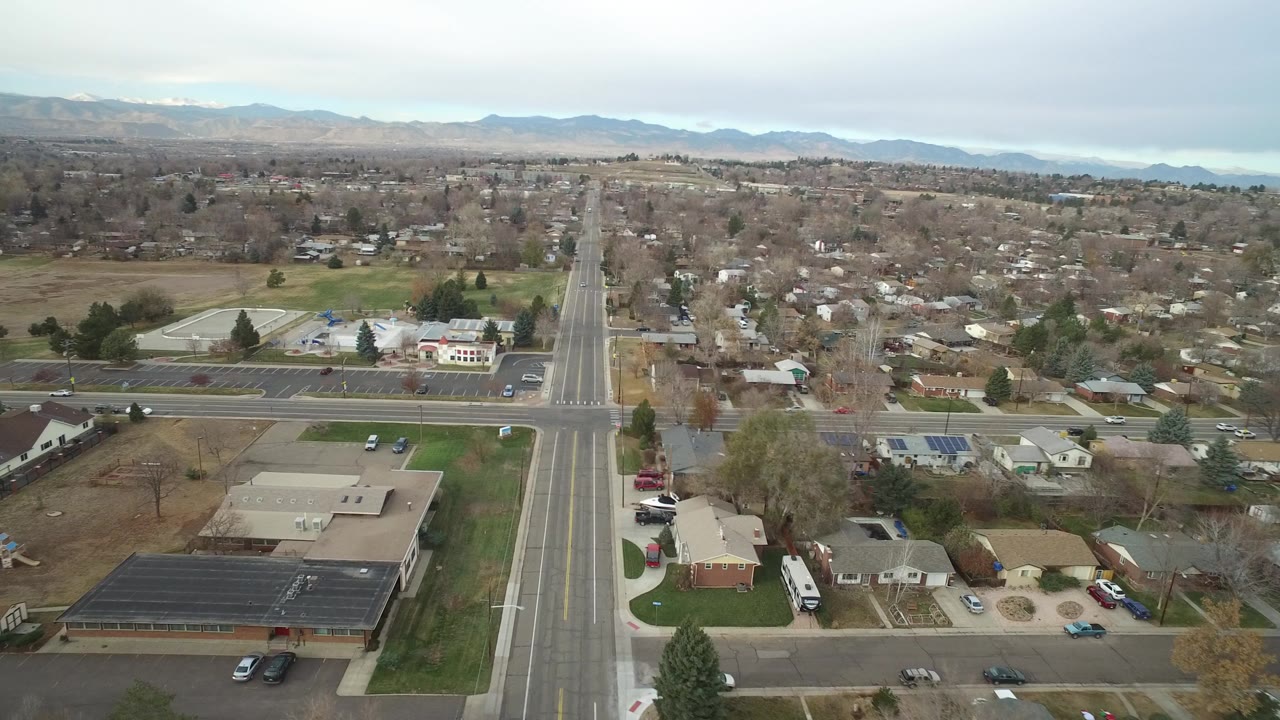 Opening aerial frame over Westminster, Colorado showing a straight road crossing a wider street, nearby homes, school grounds, parking lots, bare trees, distant mountains, and no visible people.