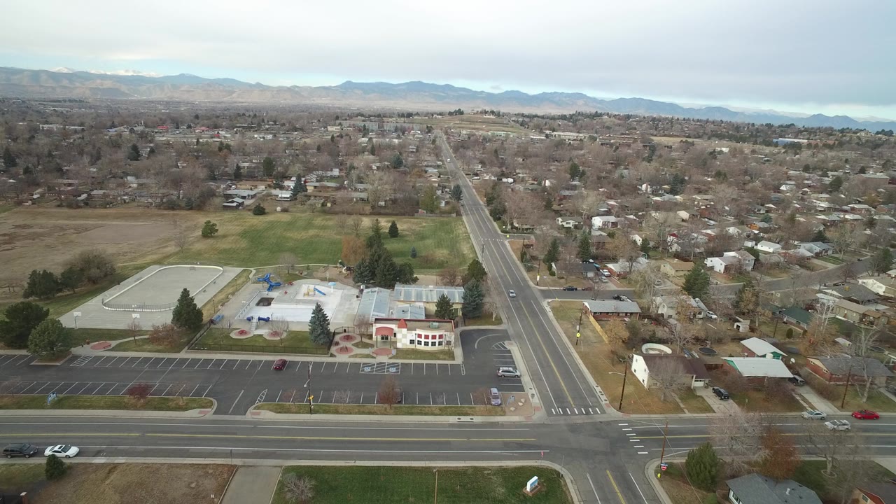 Aerial frame over Westminster, Colorado showing a road intersection, parking lot, low public buildings, surrounding homes, leafless trees, and distant mountains, with no visible people.