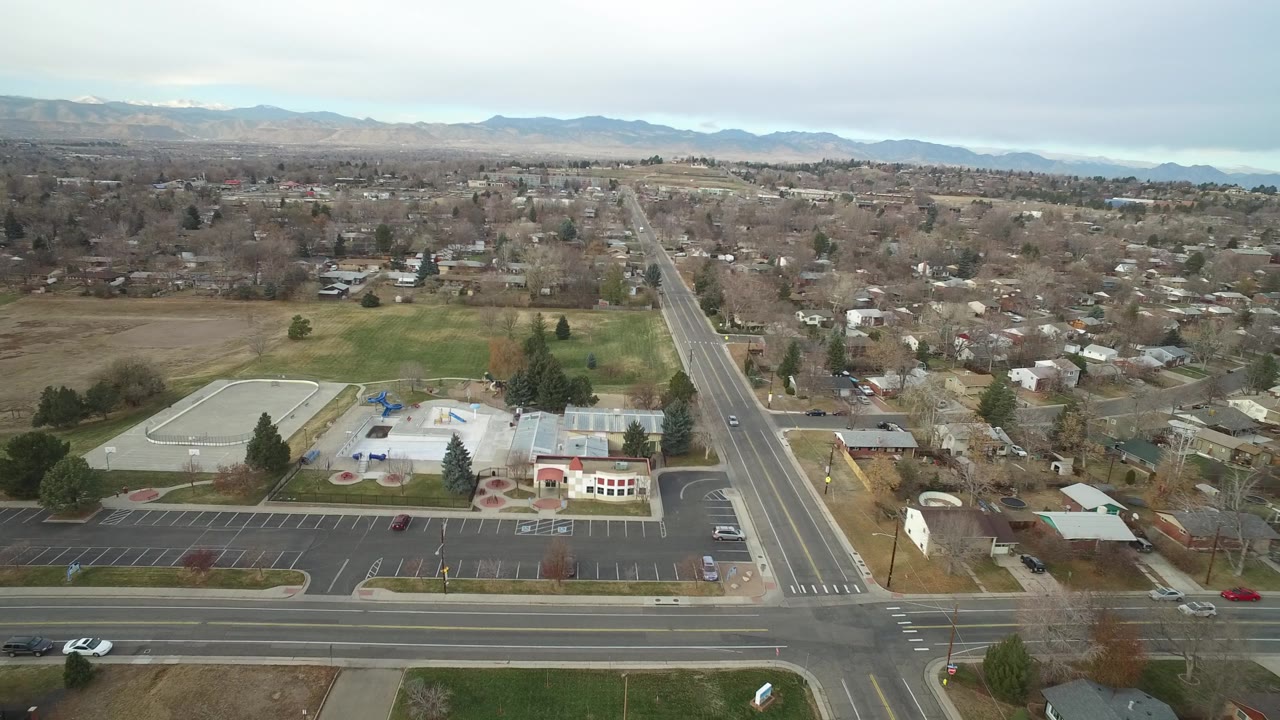 Aerial view in Westminster, Colorado with the main road centered more prominently, a civic or school complex below, open grass areas, houses, and hazy mountains in the background, with no visible people.