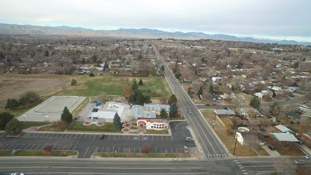 High aerial view of neighborhood blocks, a wide road, public facility grounds, and mountains.