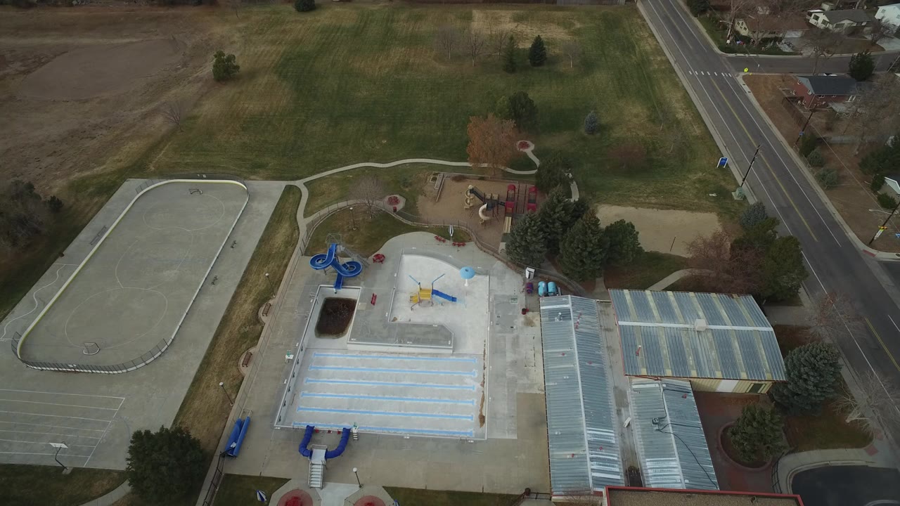 High aerial view of a Westminster, Colorado park showing a splash pad, playground, open field, road, and no visible people at the start of the clip.