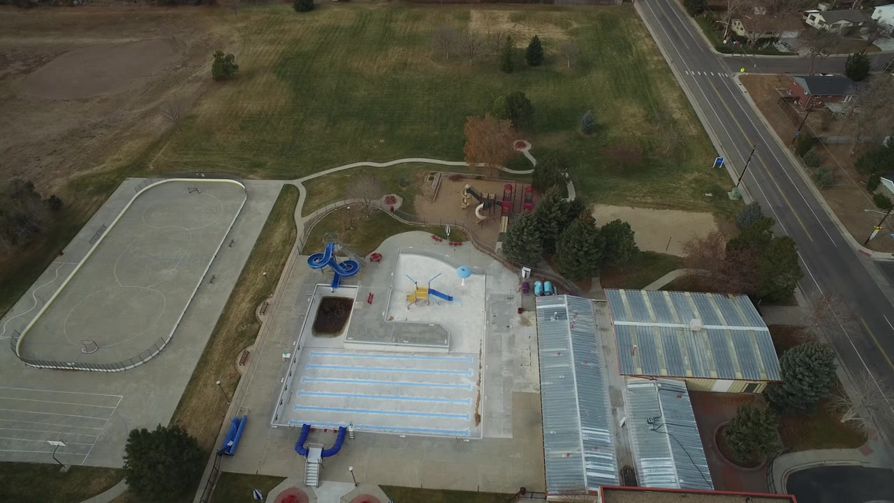 Aerial frame over Westminster, Colorado with the concrete splash pad lower center, a playground beyond it, open grass above, a road at right, and no visible people.