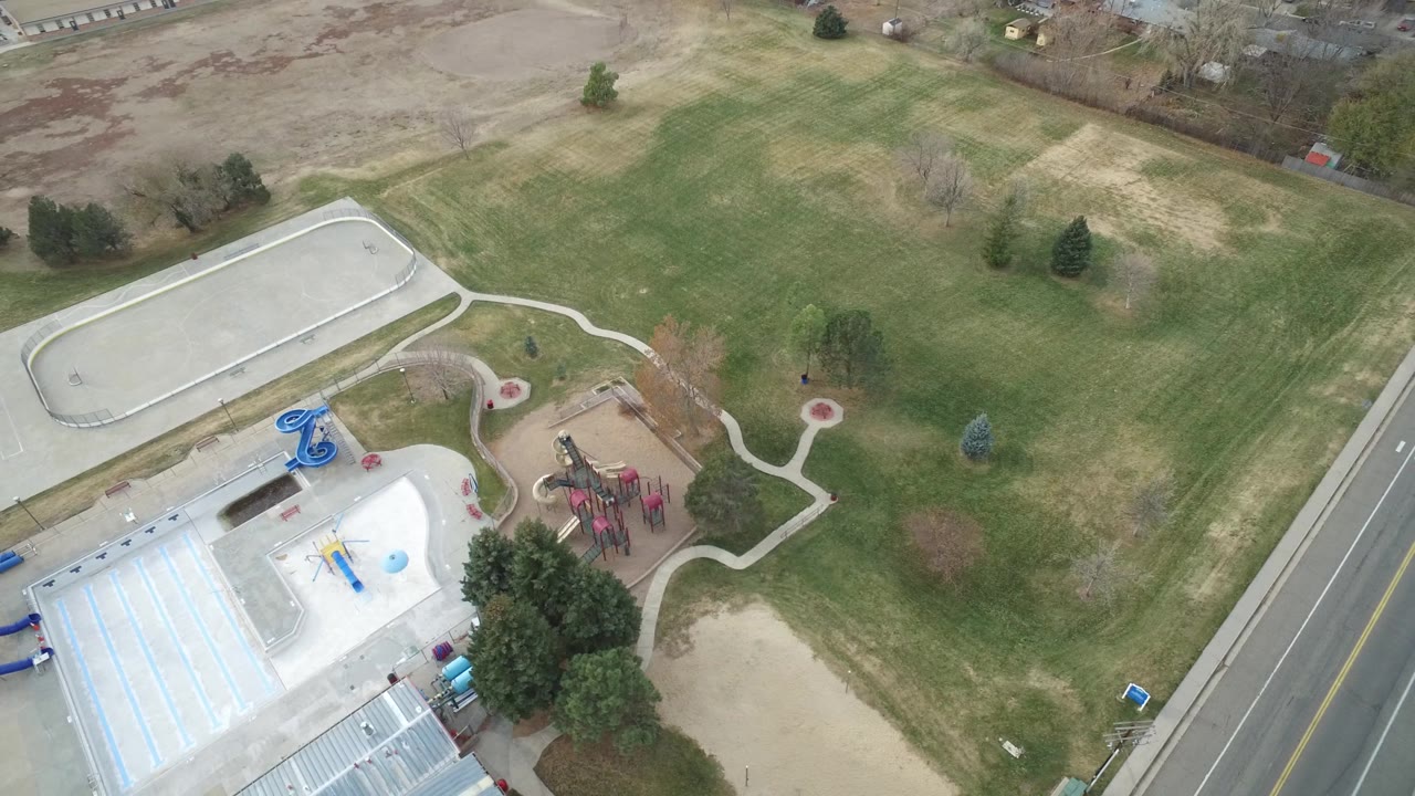 Aerial frame over Westminster, Colorado with the road more centered beside the recreation area, showing lawns, a light pool deck, blue water-play fixtures, playground equipment, paths, trees, and no visible people.