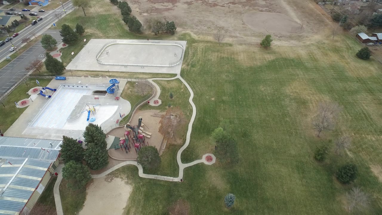 The second aerial frame keeps the Westminster park centered with the playground below the sport court, the road and houses near the top edge, and broad grass to the right.
