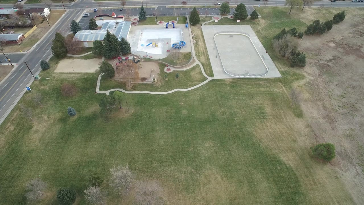 High aerial view in Westminster, Colorado showing a broad green lawn below a playground, curved walkways, a concrete recreation pad, roads, parked cars, nearby buildings, and tiny possible people.