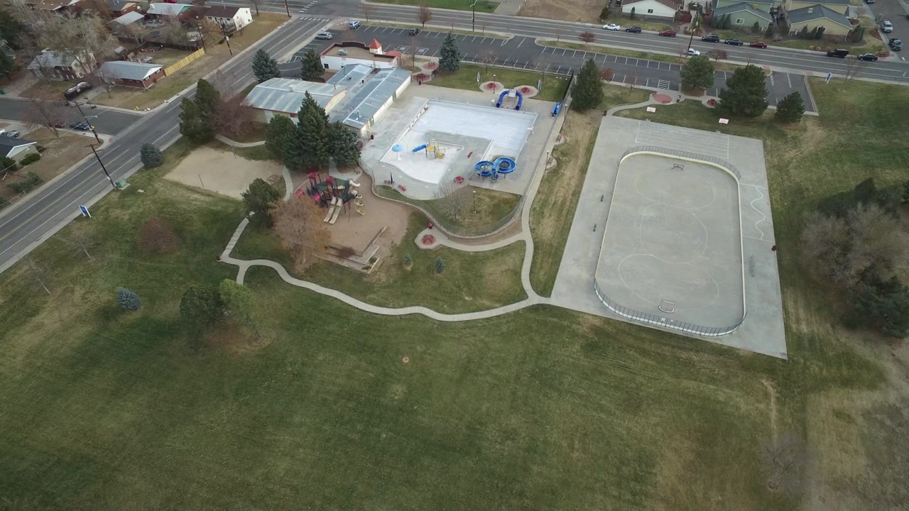 High aerial frame over Westminster, Colorado parkland with a playground, white-roofed pool area, concrete court, parking lot, road, nearby homes, and no visible people.