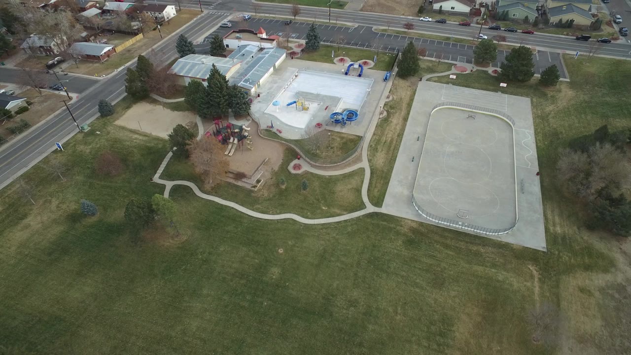 Aerial view in Westminster, Colorado with the empty concrete court more centered beside grass, the pool or recreation facility, parking rows, roads, houses, and no visible people.