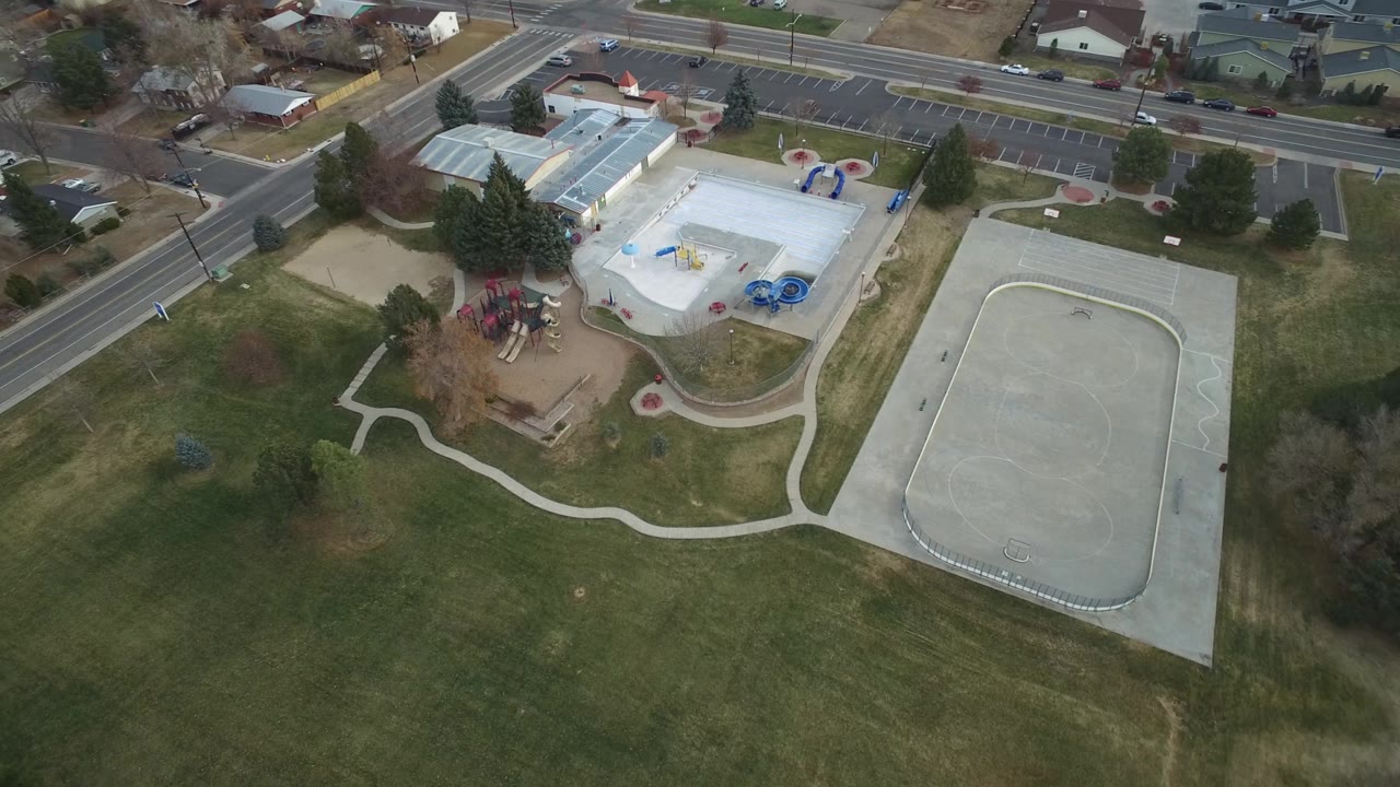 Drone view over grass, playground, pool building, concrete court, parking lot, and neighborhood streets.