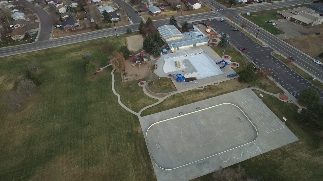 Aerial view in Westminster, Colorado with the paved court lower in frame, broad grass across the middle, and the building, parking rows, streets, and houses near the top edge.
