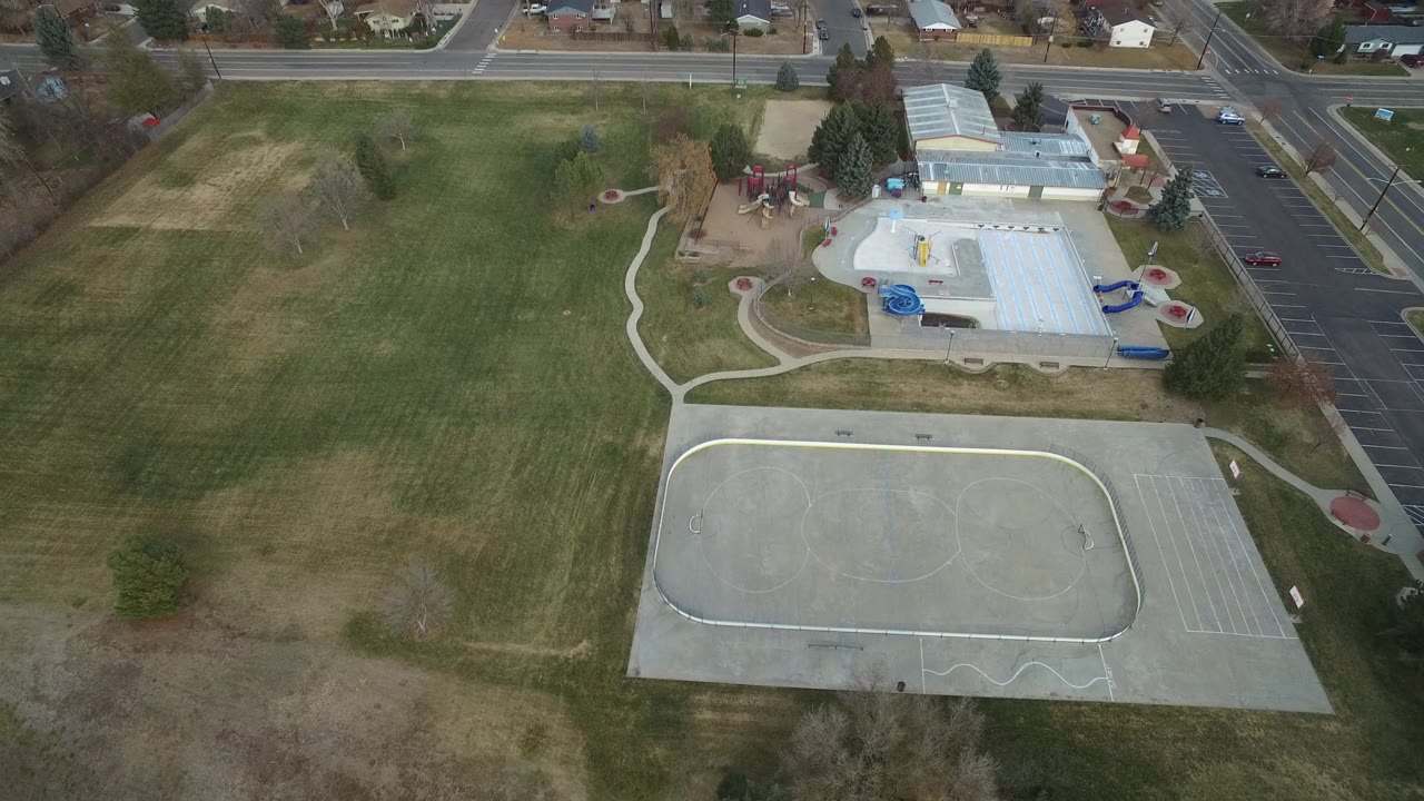 Opening overhead frame in Westminster, Colorado showing a large grass field, a concrete sports court, a recreation building with blue slide structures, surrounding roads, parked cars, nearby houses, and no visible people.