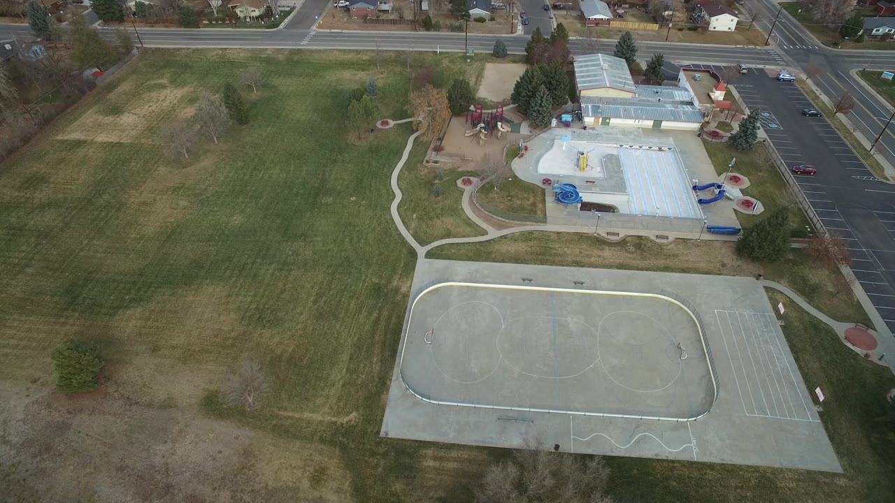 Aerial park view with field, pool-like building, sports court, parking lot, streets, and houses.