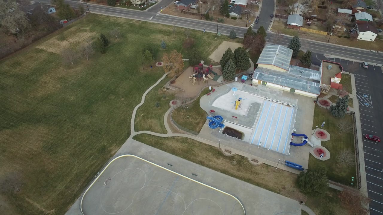 Aerial view of a pool facility, paved court, parking lot, roads, lawns, and homes.