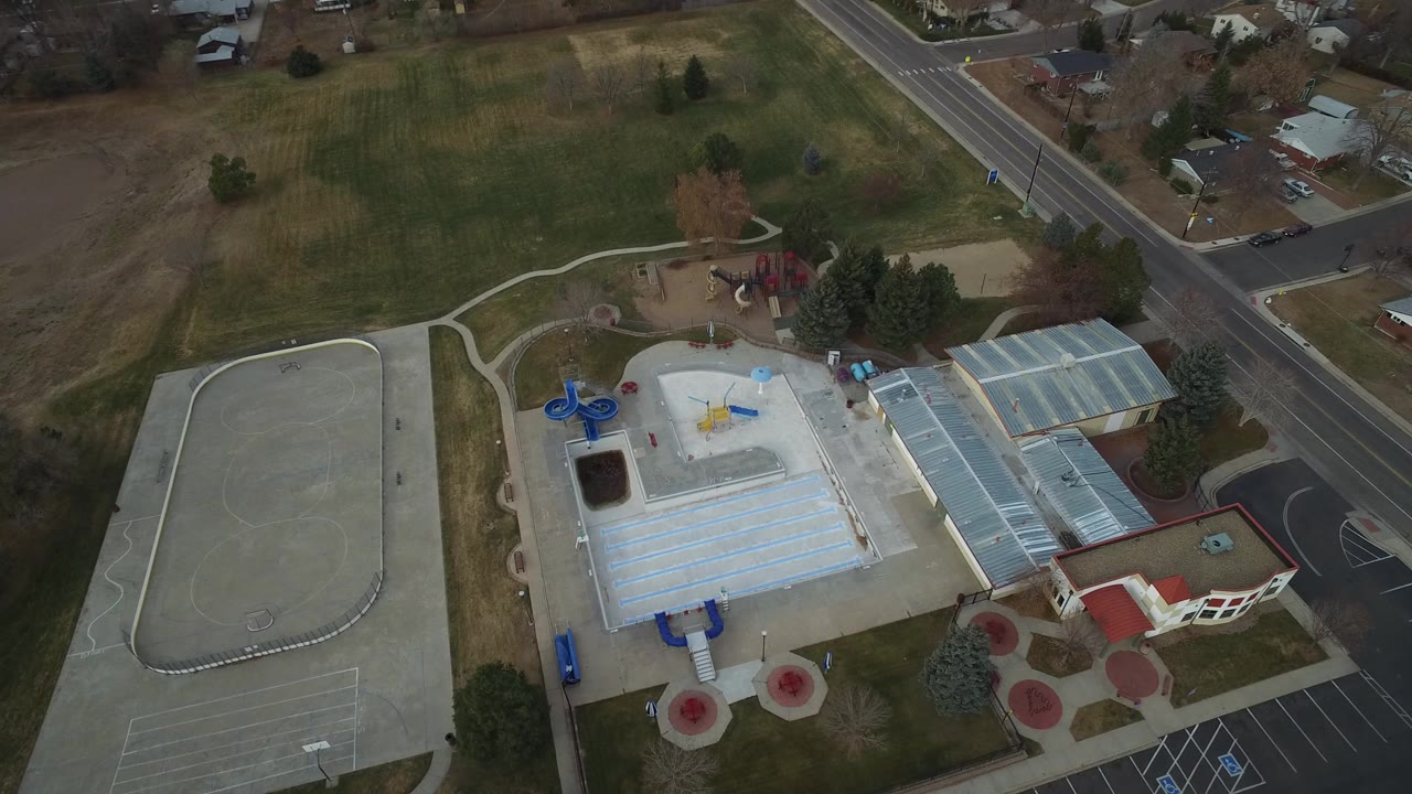Opening overhead frame in Westminster, Colorado showing a large light-roofed building, adjacent paved lot, playground area, grass fields, roads, nearby homes, and no visible people.